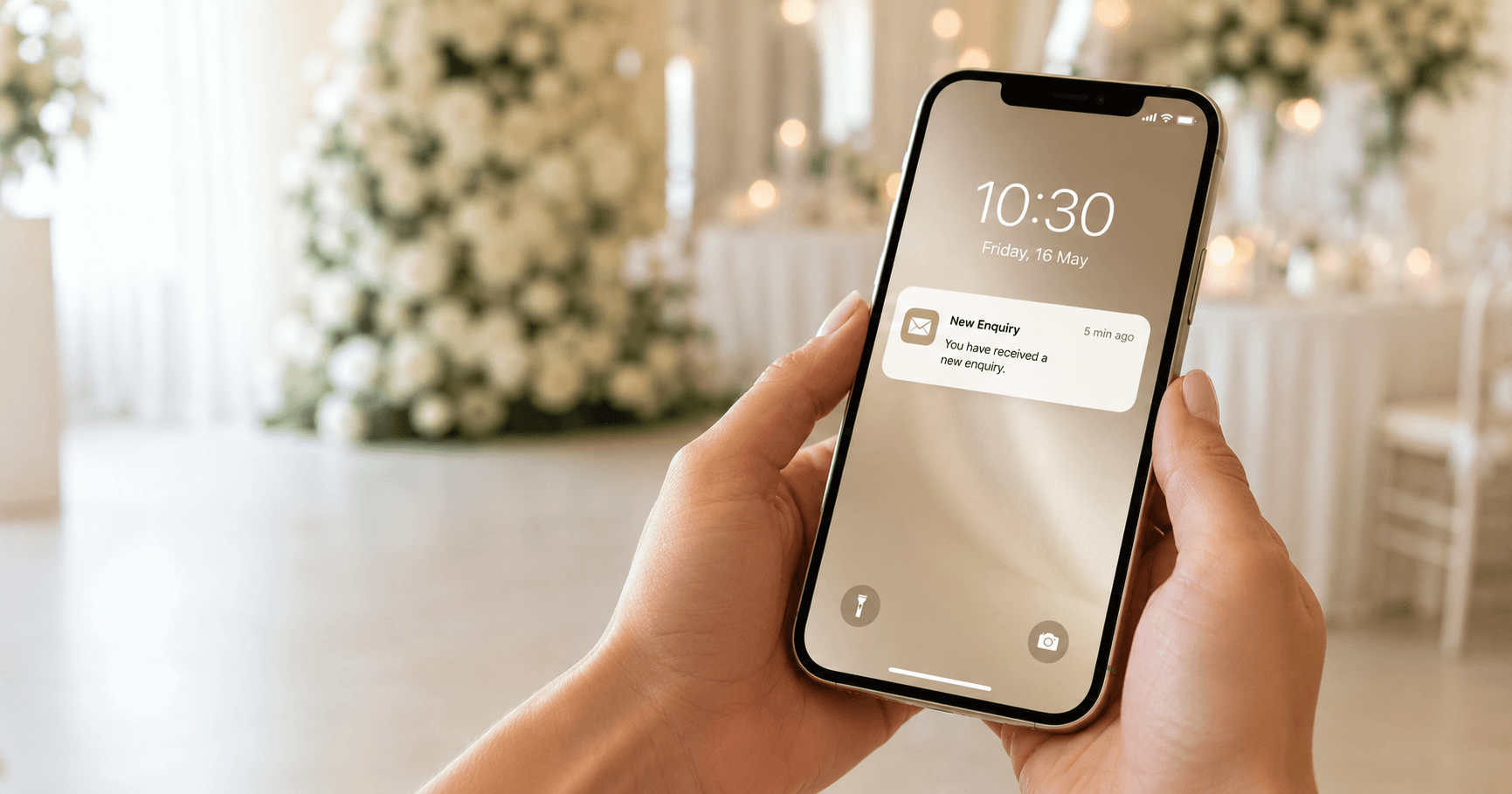 A hand holding a smartphone showing a fresh enquiry notification timestamped five minutes ago, with softly lit white wedding florals out of focus behind it.